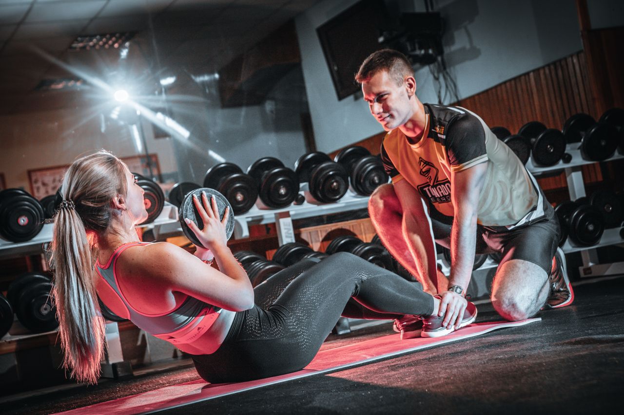 A personal trainer assists a woman doing sit-ups with a medicine ball in a gym setting.